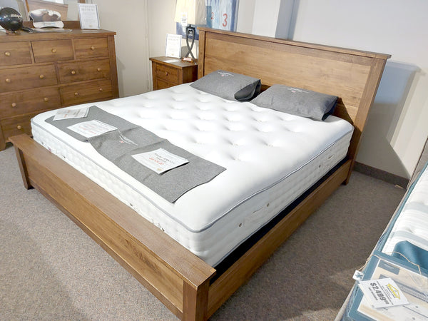 In a showroom, the Vokes 250 Rough Sawn King Bed features a single panel foot board, complemented by a white mattress, two gray pillows, and informational leaflets on top. The Provincial finish adds an elegant touch to the display.