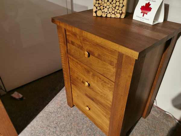 The Vokes 250 Roughsawn 3-Drawer Night Table, crafted from warmy maple with provincial finishes and featuring round knobs, is elegantly displayed on a carpet. A small log stack and a Canadian-themed card rest on top.