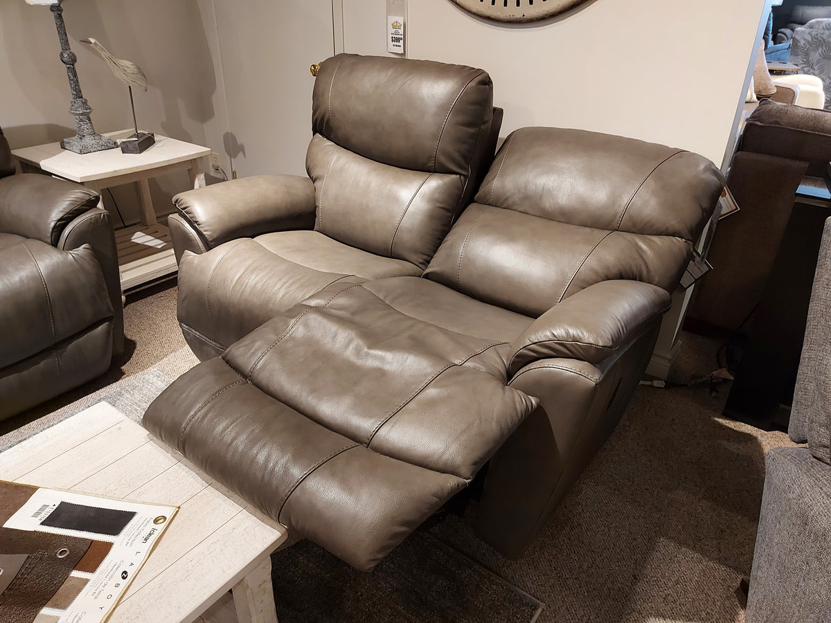 The La-z-boy 724 Trouper Leather Reclining Loveseat in gray provides ultimate reclining comfort with its split back cushions. It adds a stylish touch to any living room, enhanced by an extended footrest beside a wooden table and lamp.