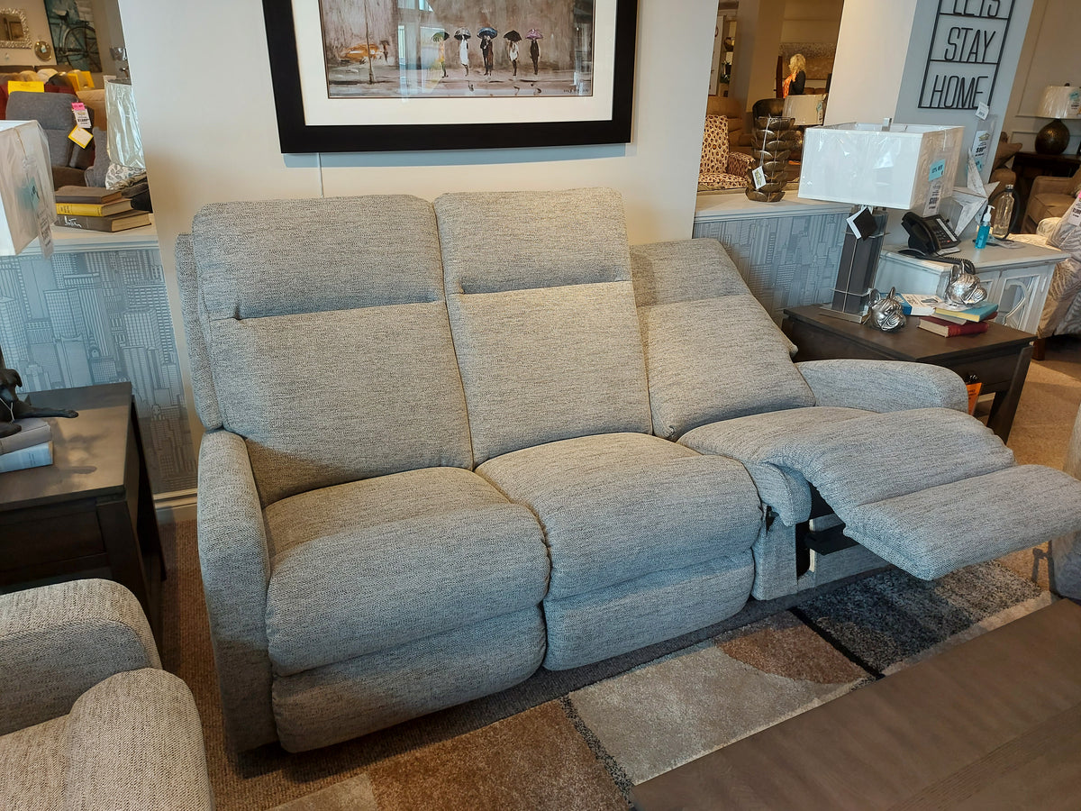 The La-Z-Boy 747 Finley Power Reclining Sofa, an ultra-plush gray masterpiece, graces the furniture store showroom with its inviting comfort and style. Surrounding tables complement this design, making it the focal point of the room.