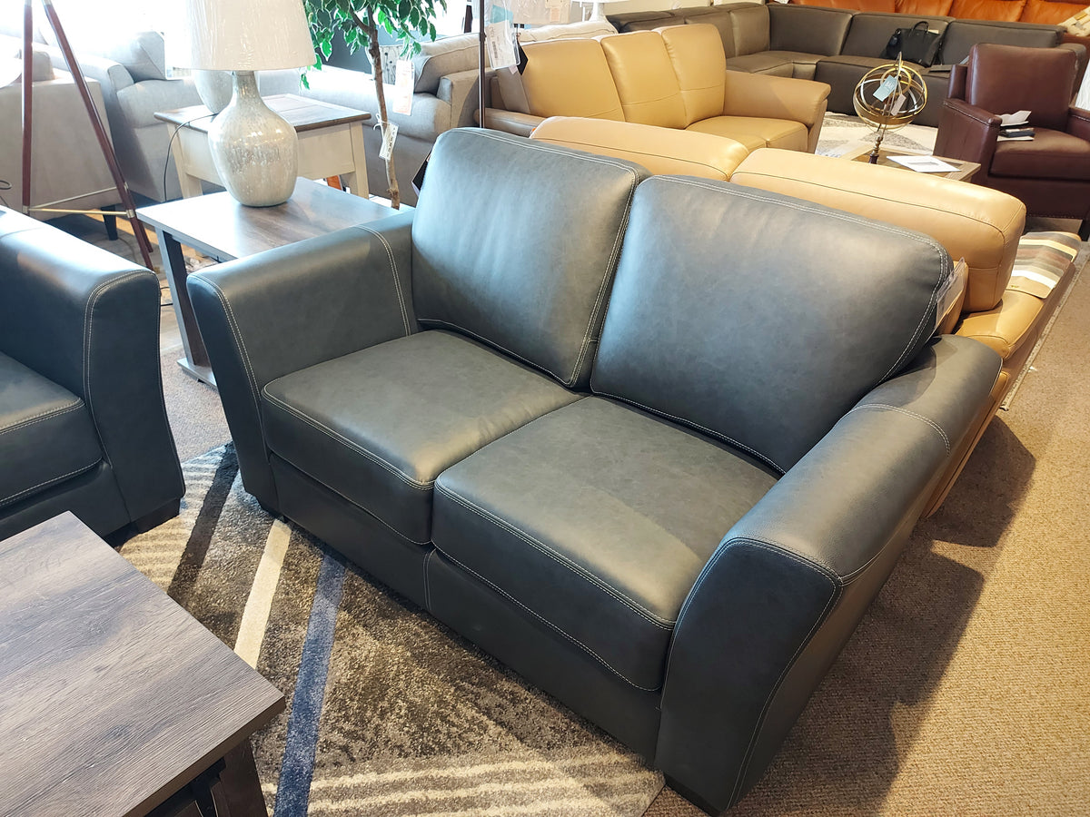 A classic gray leather loveseat from Leather Craft, called the Bayview Leather Loveseat, is showcased in a furniture store, surrounded by adaptable wooden tables and other chic sofas.