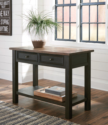 The Ashley Tyler Creek Sofa Table with drawers and a shelf sits by a window in a bright room, adorned with a plant.