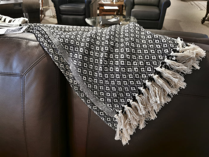 The Jete Kali Throw by Agence Viva, featuring a black and white diamond pattern and fringes, is draped over the arm of a brown leather couch.