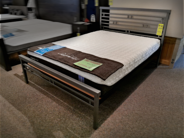 An Orson Bed Frame by Amisco, featuring a sleek metal design and complete with a mattress, is on display in the showroom with sale tags and informational labels.