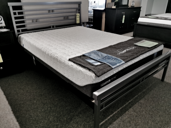 The Orson Bed Frame by Amisco showcases a sleek metal design, complemented by a white mattress and gray blanket, elegantly displayed in a store showroom.