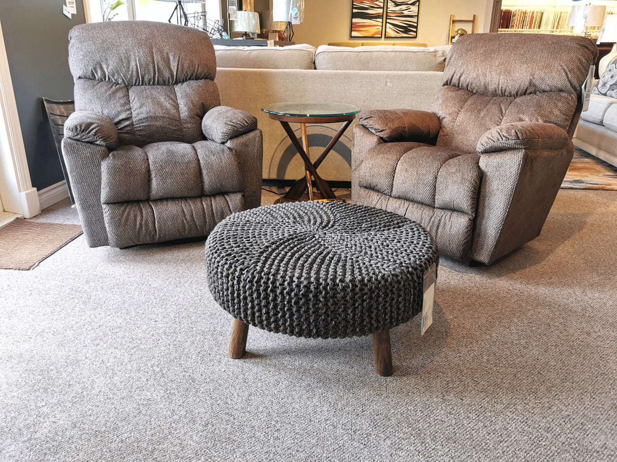 Two gray recliner chairs from Ashley Furniture flank a small wooden side table, all resting on a carpeted floor. Completing the cozy setup is the Jassmyn Ottoman by Ashley, crafted from hand-knitted yarn and adding both charm and texture to the space.