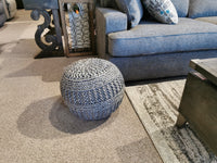 The Ashley A1000559 Benedict Pouf, in gray, is hand-knitted and rests beside a blue sofa and dark wood coffee table.