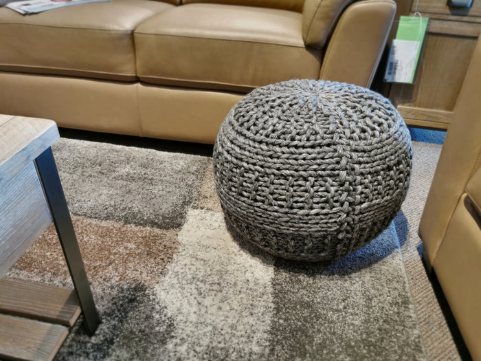 An Ashley A1000559 Benedict Pouf rests on a multicolored rug beside a leather sofa and wooden coffee table.