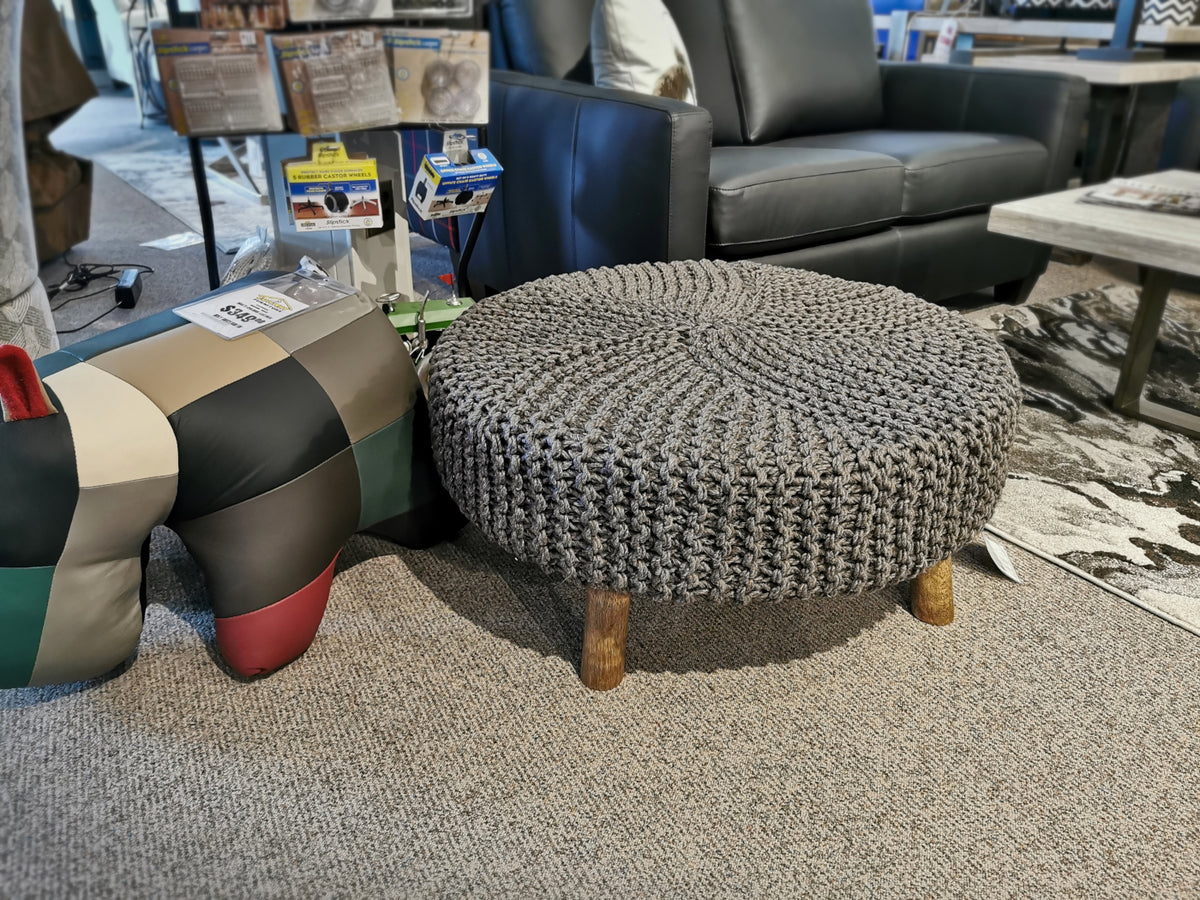 The Jassmyn Ottoman by Ashley, featuring a round design crafted from hand-knitted yarn and supported by wooden legs, is displayed alongside a patchwork-patterned cushion in an Ashley Furniture showroom.