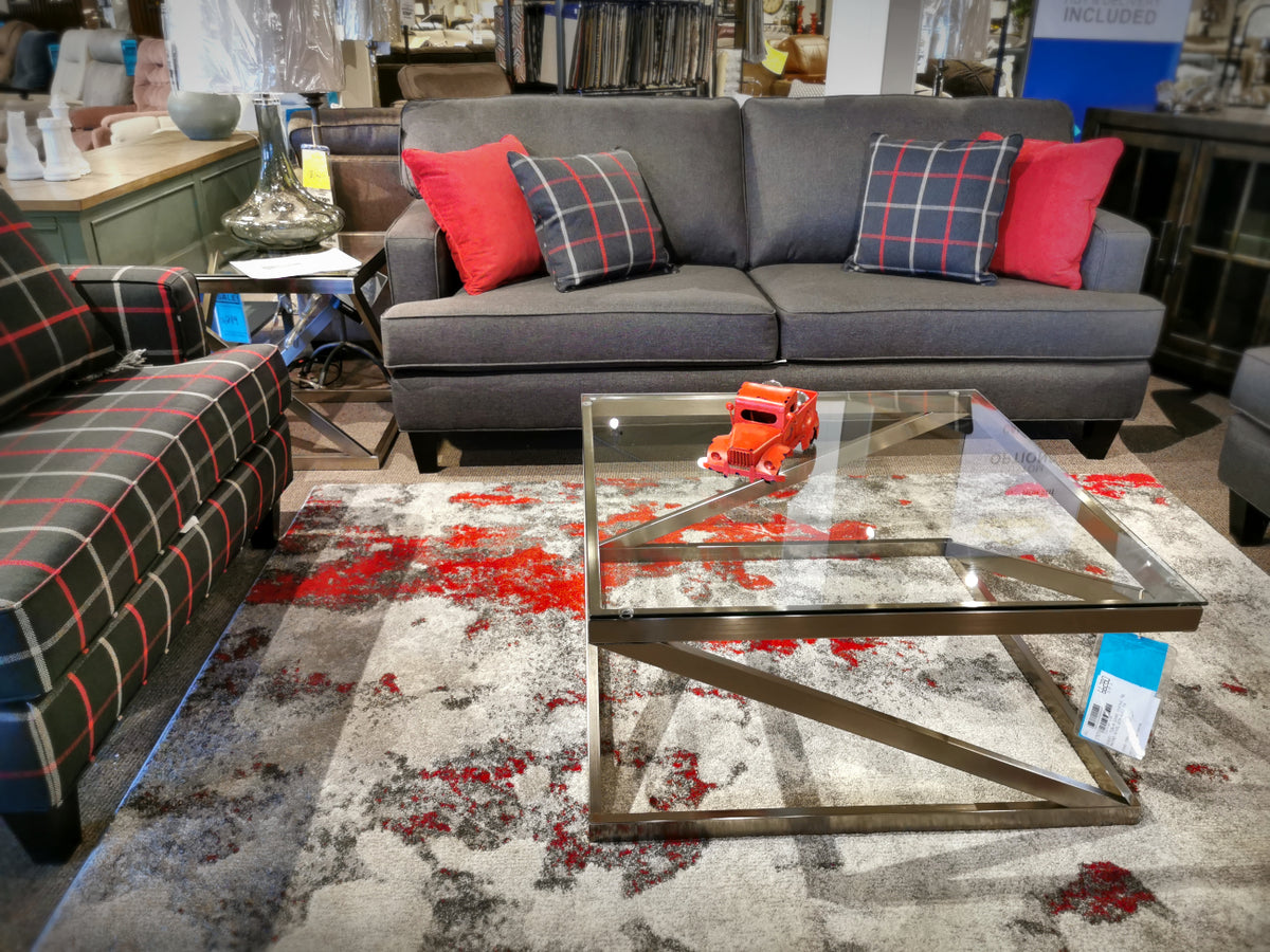 Showroom with a grey sofa, plaid armchairs, a glass coffee table, and an Ashley Coylin Square End Table on the patterned rug.