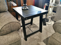 An Ashley T351 Noorbrook Table in distressed black wash with a lamp sits between two gray couches on a carpeted floor.