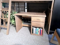 Crate's 6430 Kid's Desk in a light brown brindle finish offers a compact design with a two-drawer cabinet beneath a loft bed, making it ideal for transforming any child's bedroom. It features a side shelf for neat book storage, conveniently situated next to a small ladder amidst greenery.