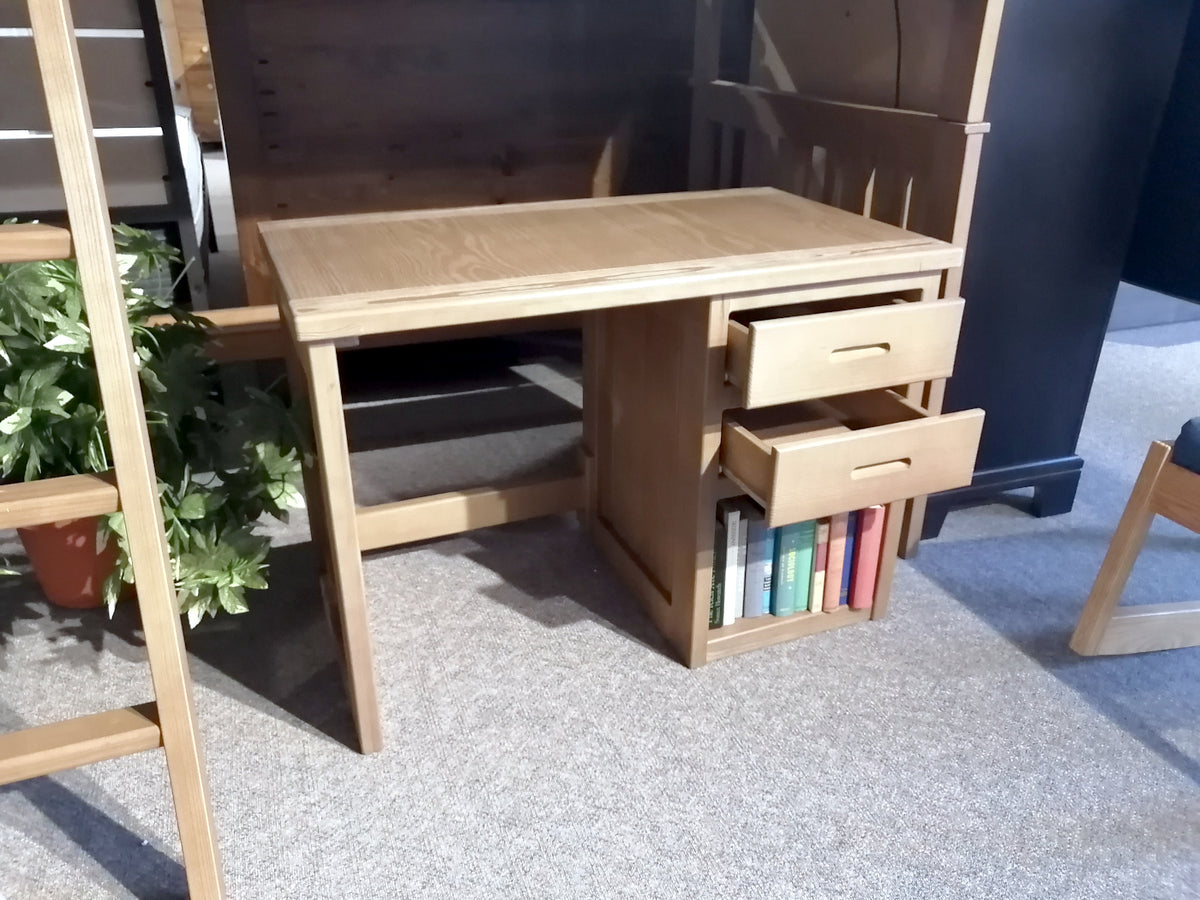 This cozy space showcases the 6430 Kid’s Desk by Crate, ideal for a child's bedroom with its light brown brindle finish. It features two open drawers and a shelf designed to hold books, complemented by a chair at the side. A plant rests on the nearby carpeted floor, adding a natural touch to the room.