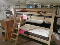 The Crate S43905 Twin Bunkbed with mattresses is showcased in a furniture store, complete with a robust ladder and complemented by wall-mounted framed art. Made from Southern Yellow Pine, this bunk bed infuses any area with rustic charm and durability.