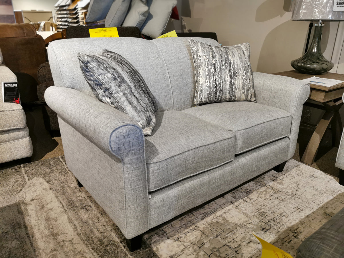 The Decor-rest 2963 Loveseat, featuring a classic design and two decorative pillows, sits on a patterned rug in a furniture showroom, making it perfect for small spaces.