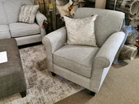 The 2963 Chair by Decor-rest, featuring a gray fabric design and a patterned cushion, sits on a textured rug and is ideal for compact spaces. It harmonizes seamlessly with a transitional sofa and matching footrest, crafting an inviting and stylish living space.