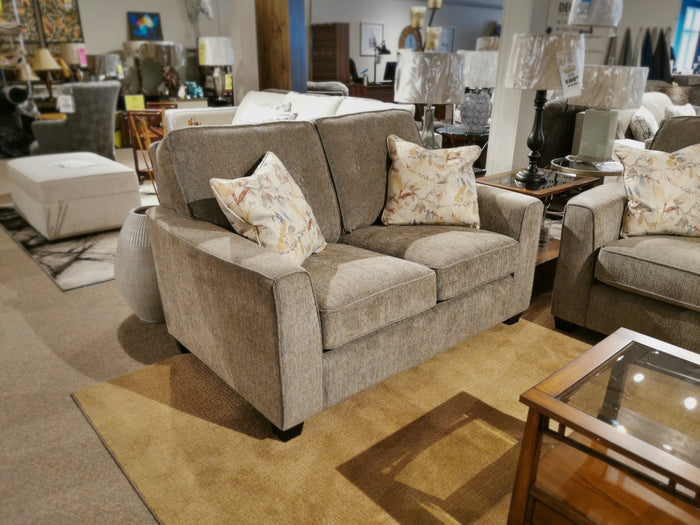 The 2967 Loveseat by Decor-rest, featuring a contemporary style and adorned with floral pillows, is showcased in a furniture showroom. Surrounding the loveseat are lamps and tables that perfectly blend elegance with functionality.