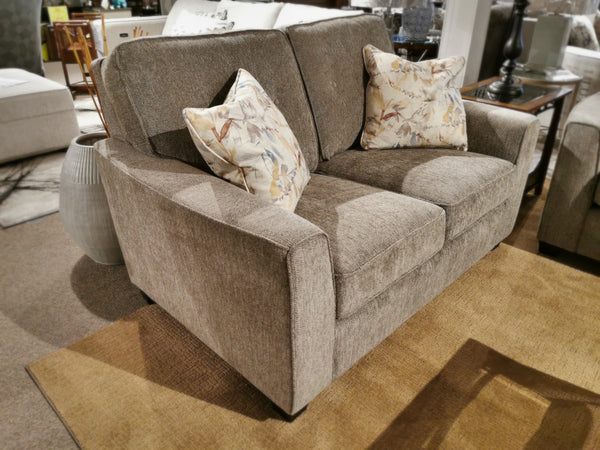 A contemporary 2967 Loveseat by Decor-rest, featuring floral-patterned pillows and customizable fabric options, is showcased in a showroom resting on a brown rug.