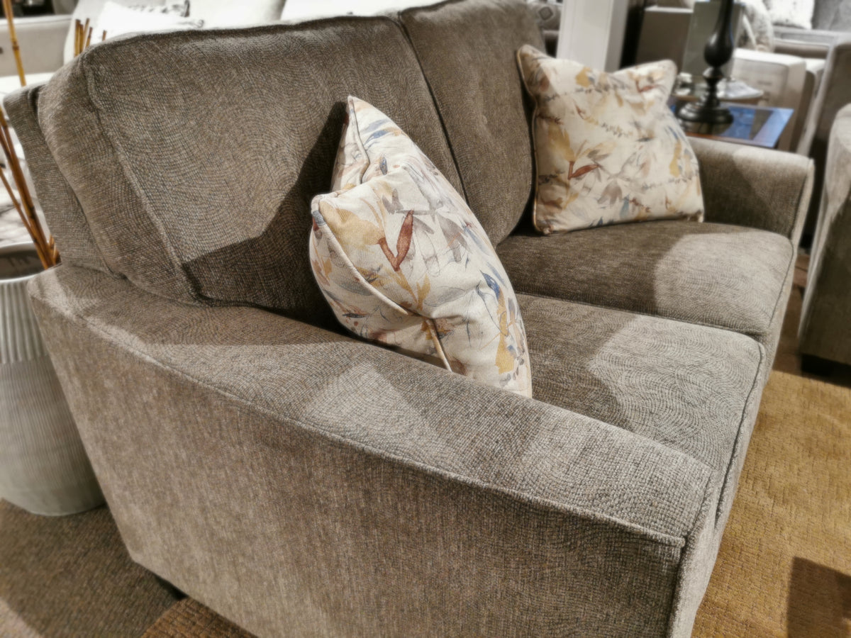 The Decor-rest 2967 Loveseat, featuring a contemporary style with two floral-patterned pillows, is elegantly displayed on a carpeted floor in the showroom, highlighting its customizable fabric options.