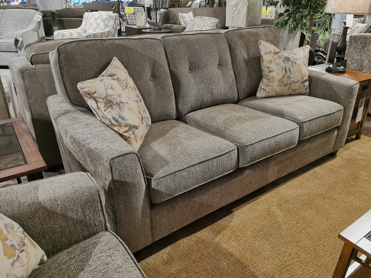 The Decor-rest 2967 Sofa, a gray three-seater with customizable fabric and floral-patterned pillows, is displayed in a furniture store with brown carpet and elegant side tables. Its contemporary style brings a modern flair to any living space.