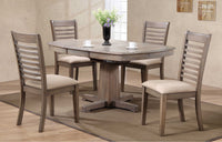 In the dining room, you'll find the elegant Ventura Dining Package by Winners Only, showcasing an oval wooden table crafted from solid hardwood with a grey washed finish. Four matching chairs surround the table, enhanced by a black vase with flowers and two cups artfully arranged on a beige rug.