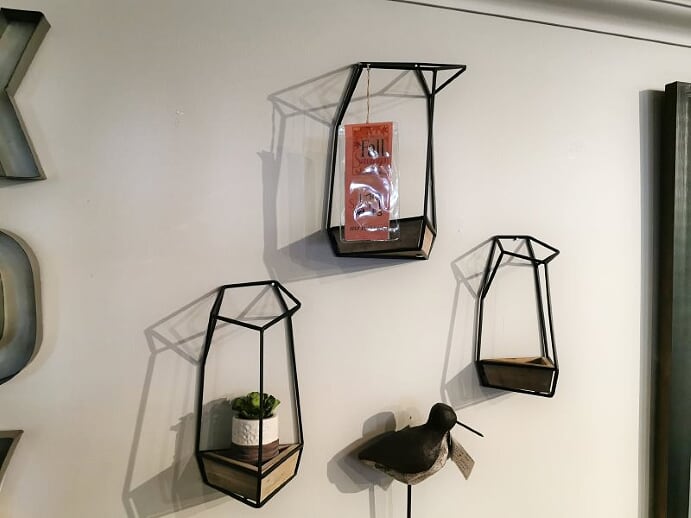 Geometric wall shelves stylishly adorn the white wall, featuring a small plant in an Imax 90426-3 Felix Wall Planter, complemented by a framed postcard and bird sculpture.