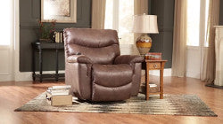 The 521 James Luxury Lift Chair from La-Z-Boy, upholstered in luxurious rich brown leather, sits gracefully on a cozy rug. It is complemented by a side table, lamp, and books while being bathed in natural light from the windows. This space-saving design enhances the living room with both style and functionality.