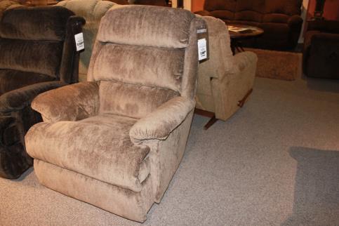 The La-Z-Boy 519 Astor Rocker Recliner, featuring its brown upholstery, is elegantly displayed in a showroom on a carpeted floor, surrounded by other exquisite furniture.