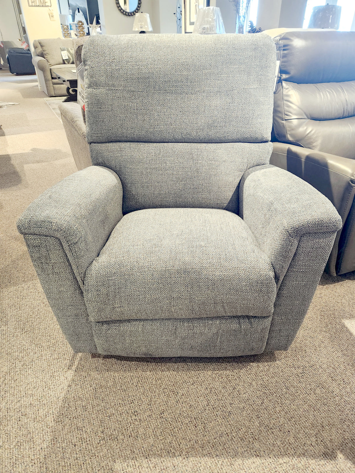 The 769 Ava Rocker Recliner by La-Z-Boy exudes comfort in a showroom setting, nestled among other furniture pieces. This gray upholstered chair offers adjustable support, seamlessly blending style and functionality.