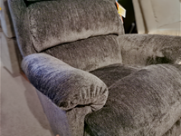 A close-up of a gray upholstered La-Z-Boy 519 Astor Rocker Recliner highlights its cushioned armrest, along with a partially visible price tag.