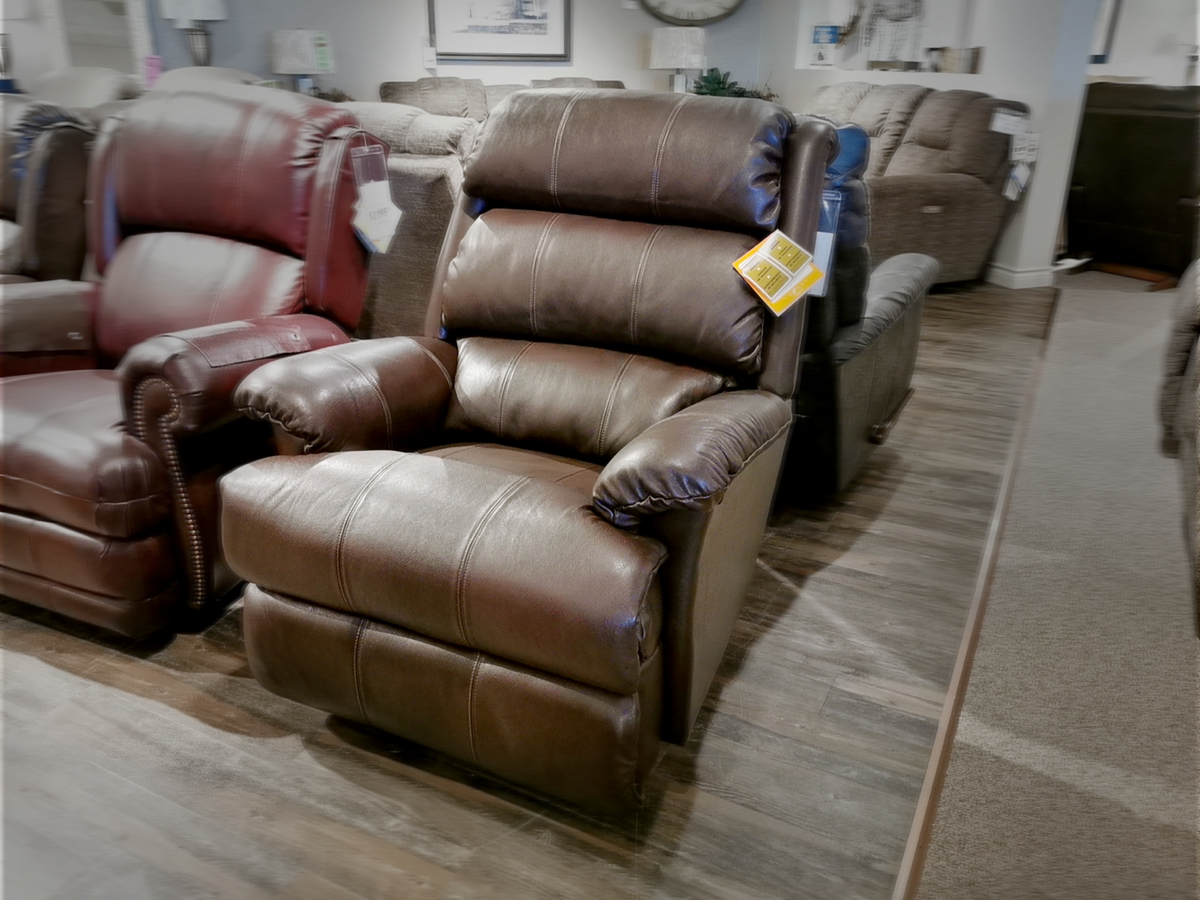 In the furniture showroom, you can find a tall brown leather La-z-boy 519 Astor Rocker Recliner, with an assortment of other stylish chairs in the background.