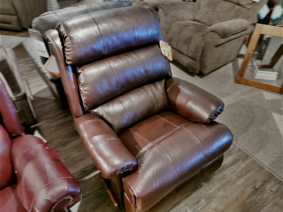 The La-z-boy 519 Astor Rocker Recliner takes center stage in the showroom, crafted from luxurious brown leather. Amidst other elegant furniture, this tall recliner ensures both comfort and style for any living area.