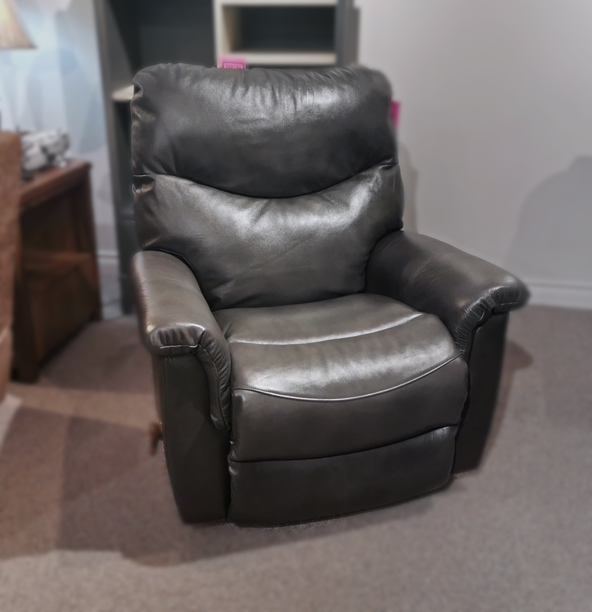 A 521 James Rocker Recliner by La-z-boy, featuring a black leather finish and a bucket-style seat, is placed in a room with carpeted flooring, surrounded by wooden furniture and a lamp.