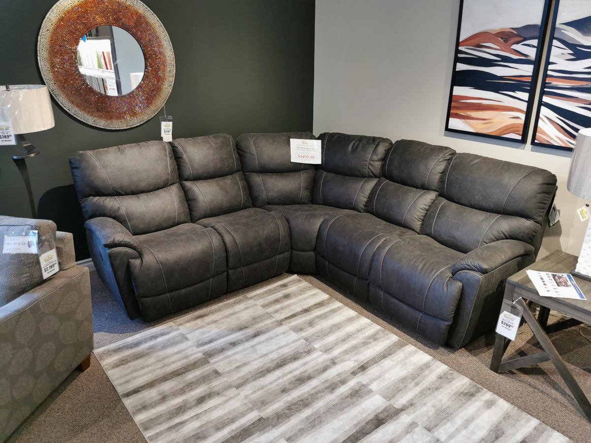 The La-Z-Boy 724 Trouper Reclining Corner Sectional in gray, featuring bucket seats and customizable fabrics, is displayed in a showroom. It is elegantly positioned on a striped rug nearby two lamps and decorative wall art.