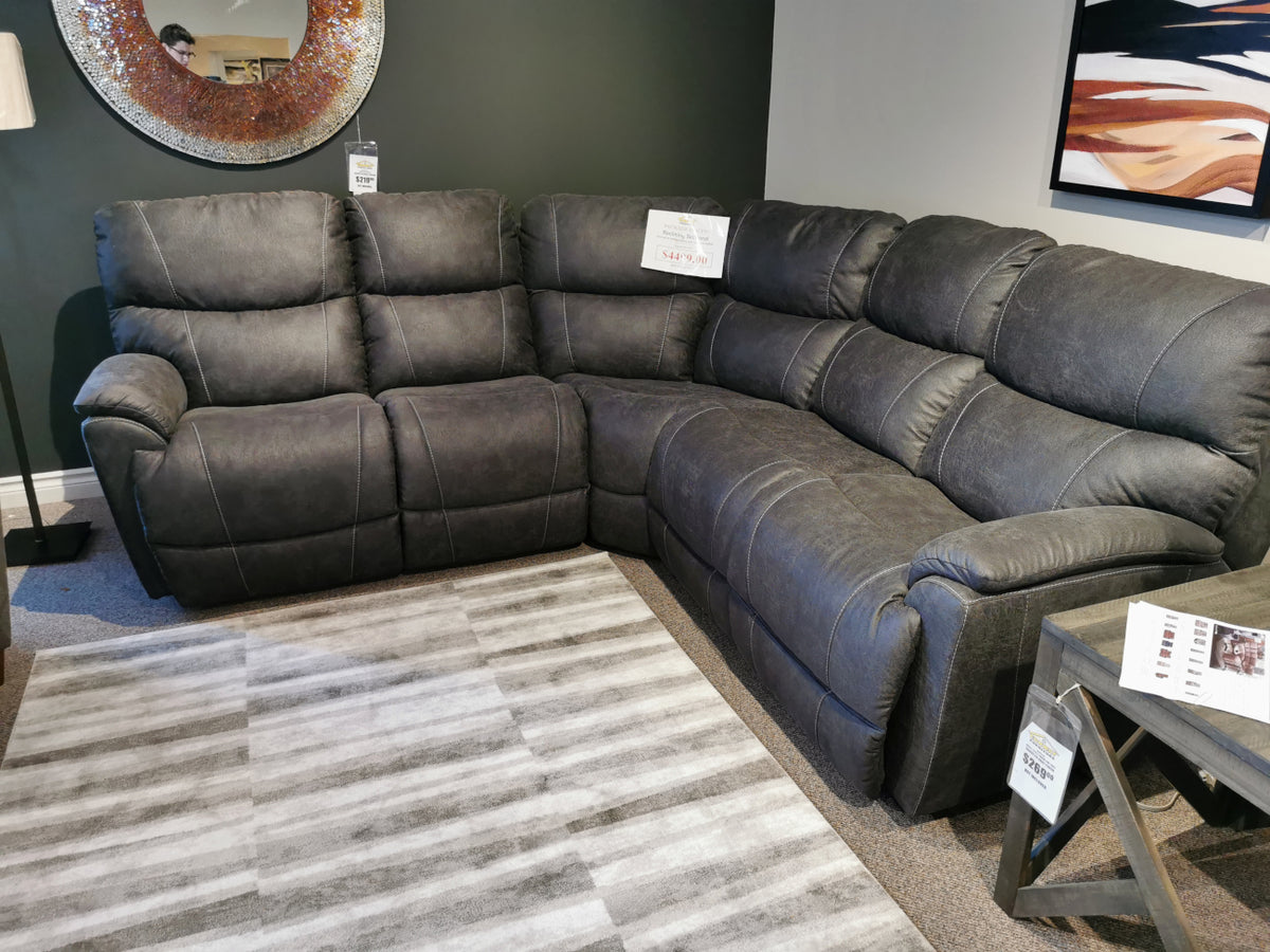 La-z-boy's 724 Trouper Reclining Corner Sectional in dark gray, showcased on a striped rug in a showroom, complemented by a round mirror and wall art.