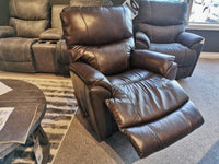 In the showroom, the La-Z-Boy 724 Trouper Leather Rocker Recliner in brown provides ergonomic lumbar support with an extended footrest, nestled among other recliners and a stylish table.