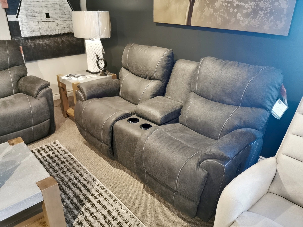 A 724 Trouper Reclining Loveseat with Console by La-Z-Boy, featuring bucket seats in a gray finish, is showcased in a living room showroom alongside a lamp and a patterned rug.