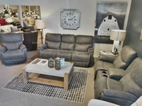 La-z-boy showroom spotlighting the 724 Trouper Reclining Loveseat with Console in gray, ideal for holding drinks or remotes. Paired with a chic coffee table and stylish lamps, and accented by a large wall clock and barn artwork to complete the ambiance.