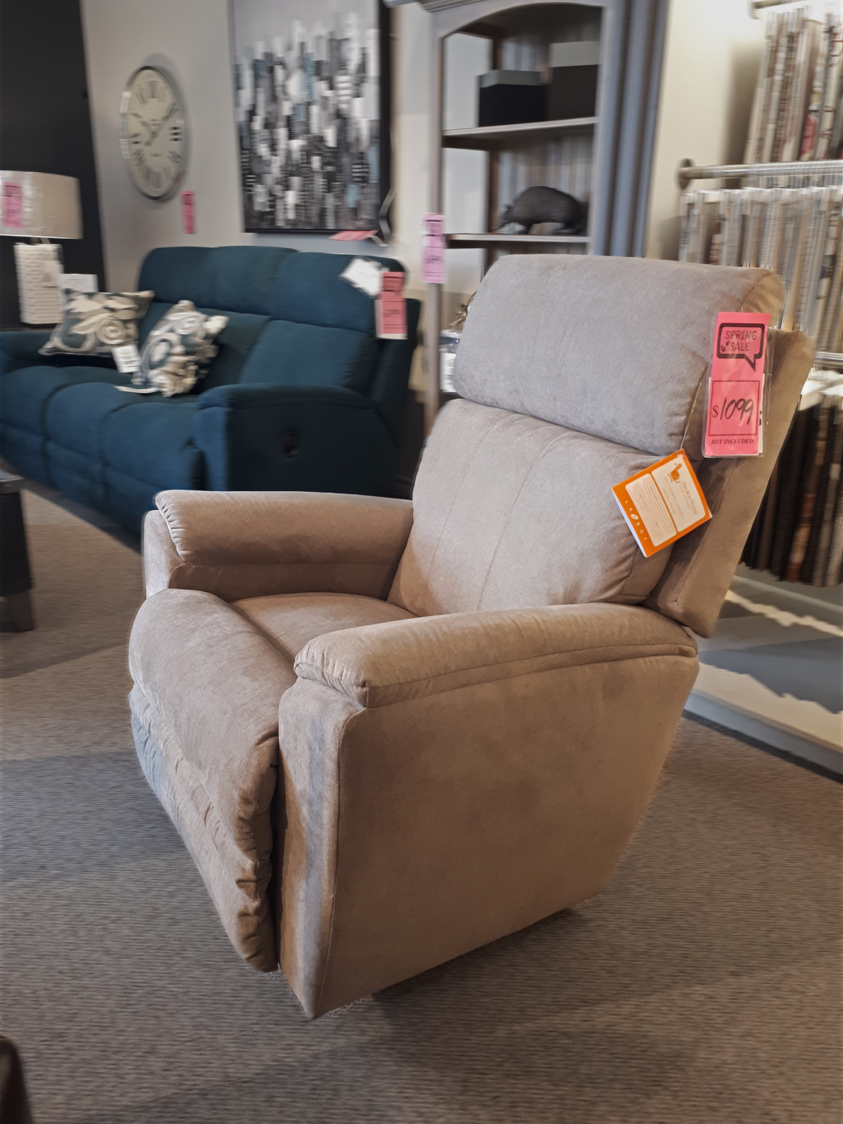 The La-z-boy 754 Talladega Rocker Recliner in beige, adorned with sale tags, is showcased in a furniture store and features a convenient USB port for modern living. In the background, a blue sofa and other elegant furnishings complement the scene.