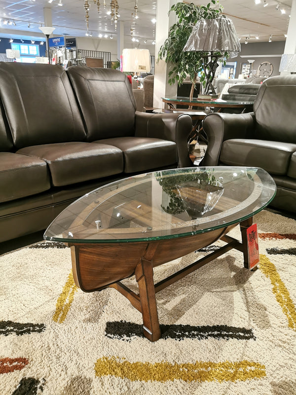 T2214 Beaufort Boat Cocktail Table by Magnussen on a patterned rug, flanked by brown leather sofas in a showroom.