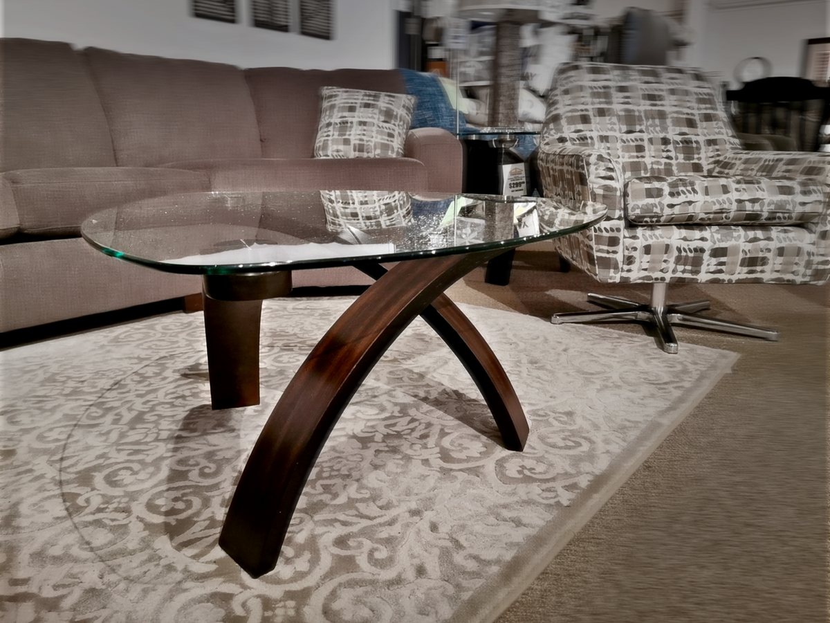 Modern living room featuring the Magnussen T 1396 Allure Glass Coffee Table—a curved glass table with walnut veneers—paired with a gray sofa and patterned armchair on a decorative rug, creating a contemporary look.