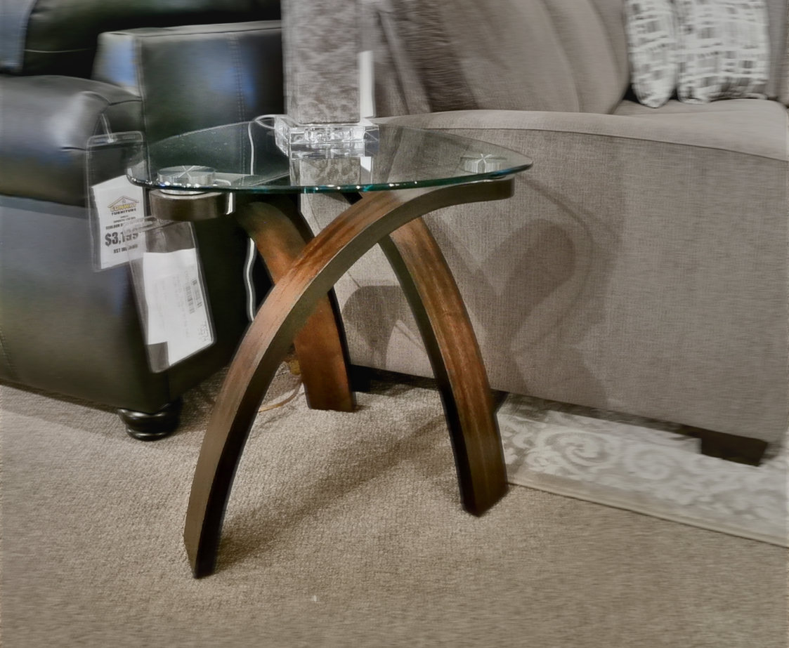 The Magnussen T 1396 Allure Glass End Table, boasting modern sophistication and curved wooden legs with walnut veneers, is elegantly positioned between a black leather chair and a gray sofa in the showroom.