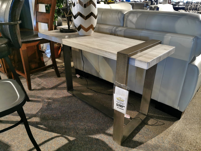 The 4701 Wiltshire Sofa Table by Magnussen, featuring a Sea Shell marble top and a Champagne Bronze metal frame, is showcased in a furniture showroom. This elegant piece, priced at $799.99, effortlessly complements contemporary upholstered settings and adds a touch of sophistication to any space.