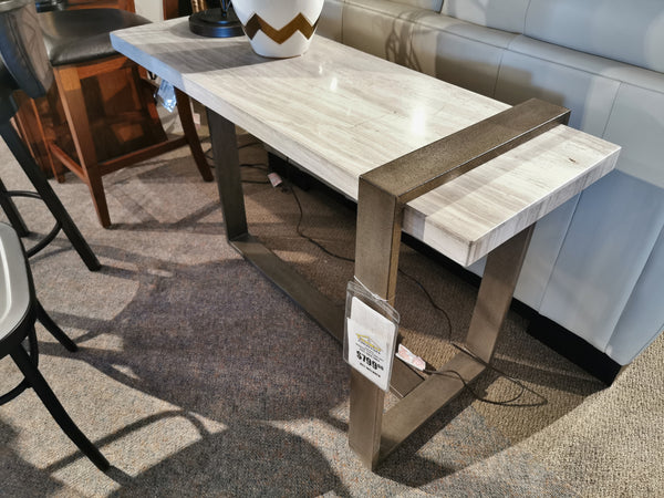 In a showroom featuring contemporary upholstered settings, the 4701 Wiltshire Sofa Table by Magnussen is displayed. It showcases a modern rectangular design with a light wood top and Champagne Bronze metal legs, with its price tag attached.