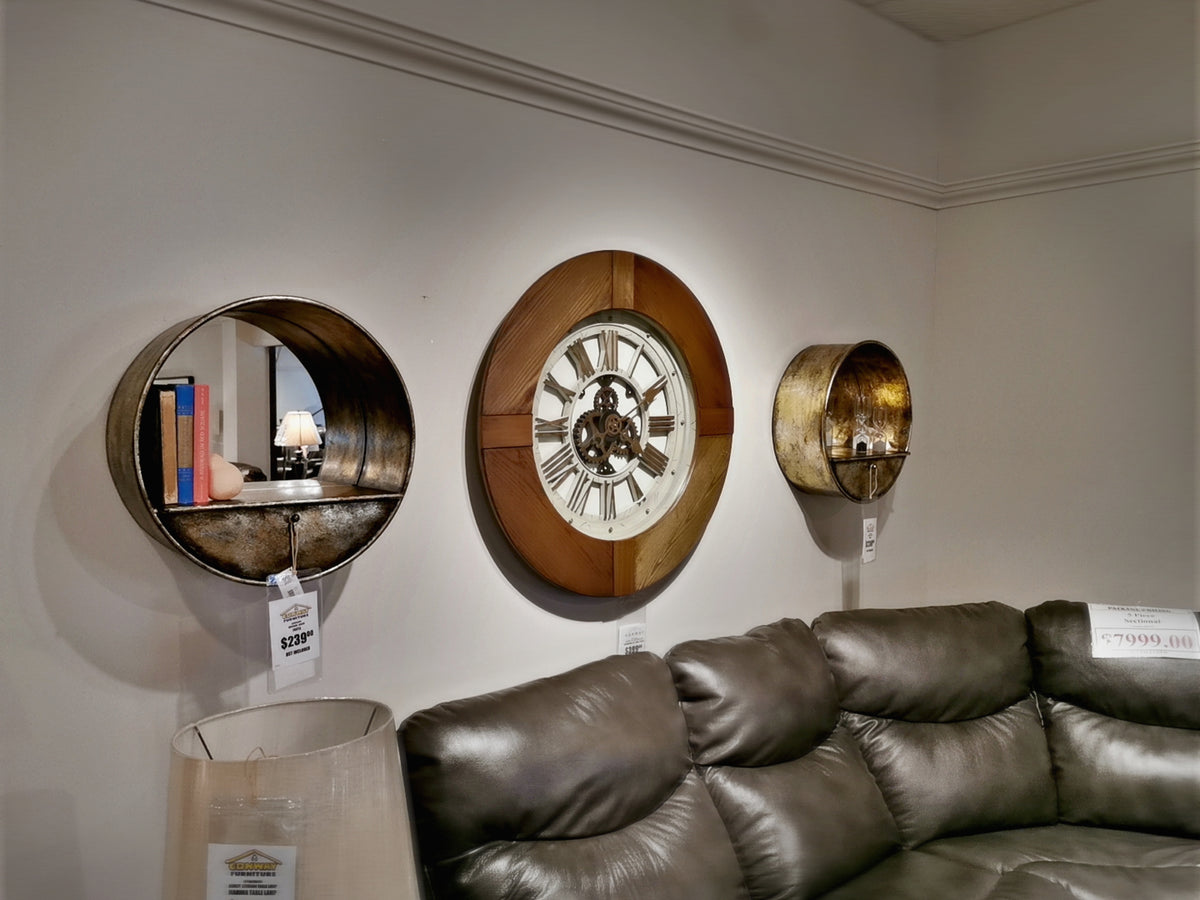 A deep-set metal mirror and the 50086 Frota Wall Shelf by Mercana complement the clock above a gray leather sofa in a furniture store display.