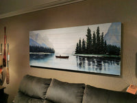 Oil painting titled "68352 Solitude" by Mercana, depicting a canoe on a lake with trees and mountains, adorning the wall above a dark sofa on a beige backdrop next to nature-inspired decorative paddles.