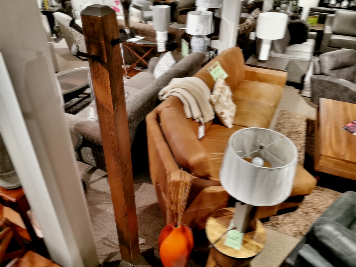 Step into our furniture showroom featuring a tan leather sofa and a wooden coffee table in an elegant Dark Hazel finish. A Munros Pine Coat Tree in Dark Hazel finish stands gracefully by the floor lamp, while cozy pillows and a throw blanket add charm to the decor.