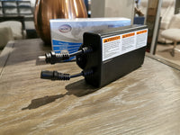 A Black rectangular power adapter with multiple cables and warning labels sits on a wooden surface, ready to charge the 49001 Battery Pack by Palliser for your Conway power recliners.