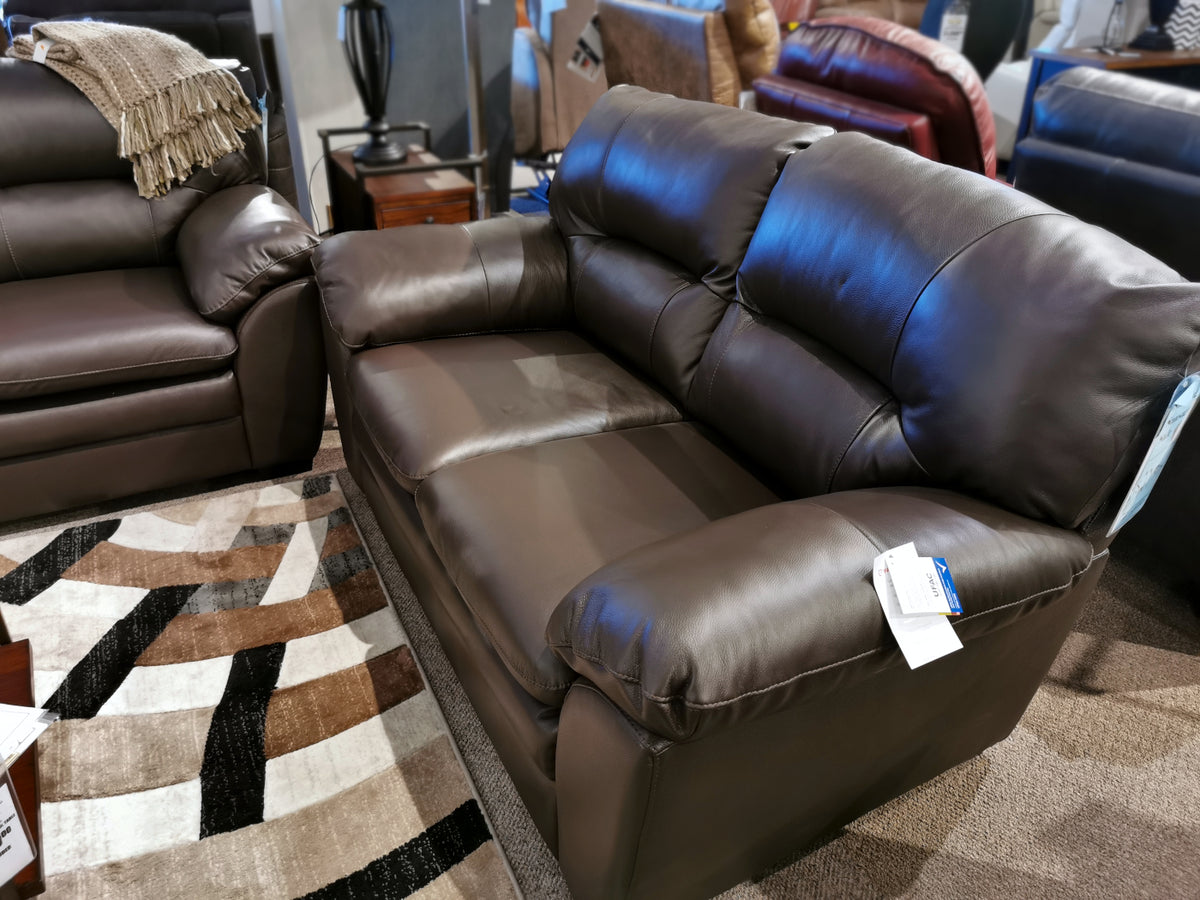 The Palliser 77343 Amisk Loveseat, upholstered in rich dark brown leather and equipped with luxuriously soft back cushions, is displayed on a patterned area rug in a showroom, providing cozy seating for any setting.