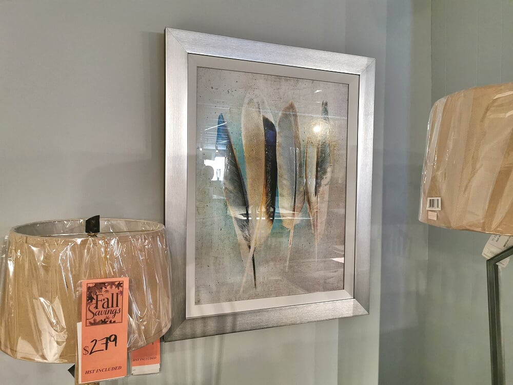 Displayed on the wall is a framed artwork titled "Turquoise Feathers" by Picture Depot in a 30" x 37" Glass Art Frame, alongside plastic-wrapped lampshades marked with a sale price of $279.